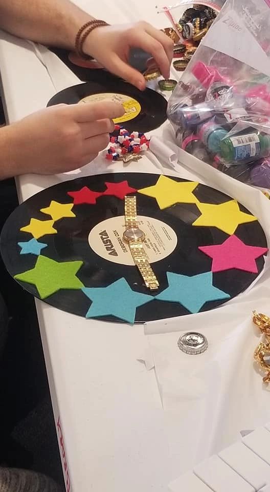 A record decorated with felt star cutouts in yellow, red, blue and green, a gold watch pressed into the label centre, an artist selecting bottle caps nearby.