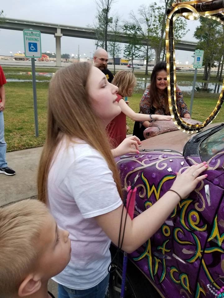An artist runs a hand over the textured painted surface of the Women Rock Art Car's hood, decorated with swirling gold script, while others look on nearby.