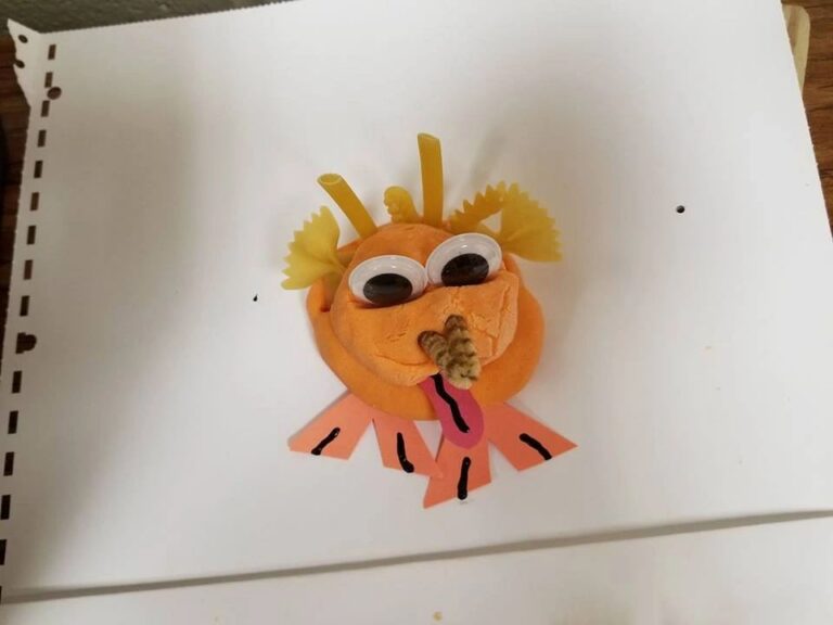 A small clay face with pasta-shaped ears, googly eyes and a pipe-cleaner tongue, decorated with paper hands and feet.