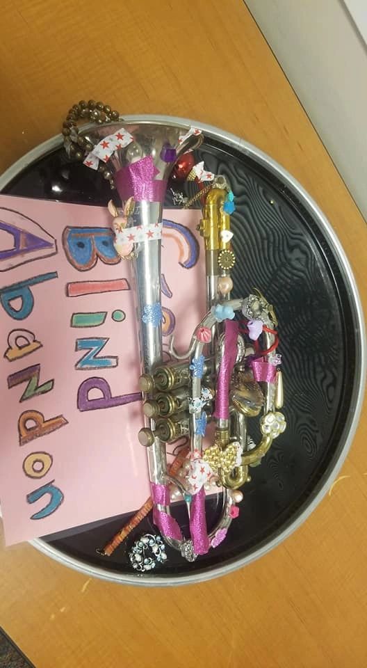 A trumpet decorated with pink glitter tape, charms and small trinkets, resting inside a black display tray beside a card that reads Creating With Blind Abandon.