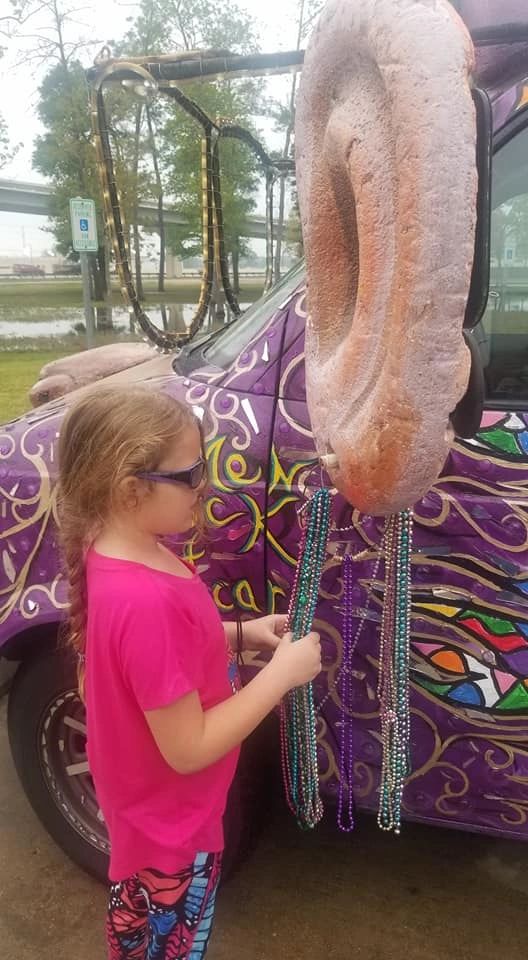 An artist holds strands of beads hanging from the sculpted ear on the Women Rock Art Car, colourful painted swirls visible on the hood.