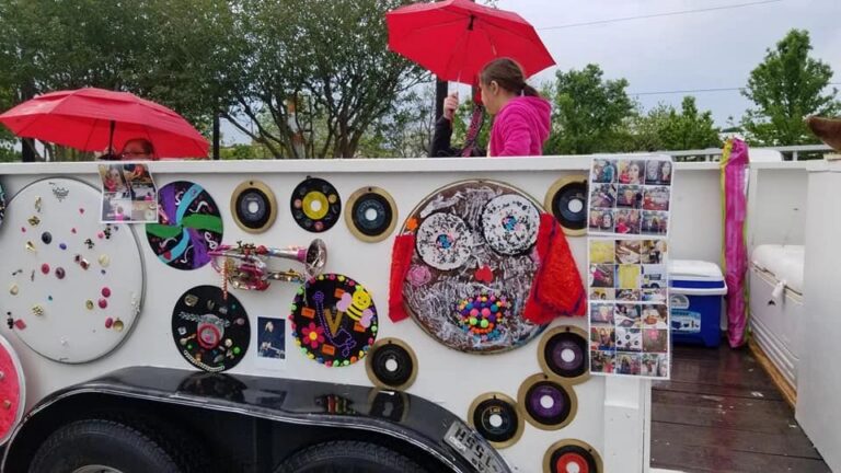 A group holds red umbrellas over the float's decorated panel of records, a trumpet and photo collages, rain visible in the scene.