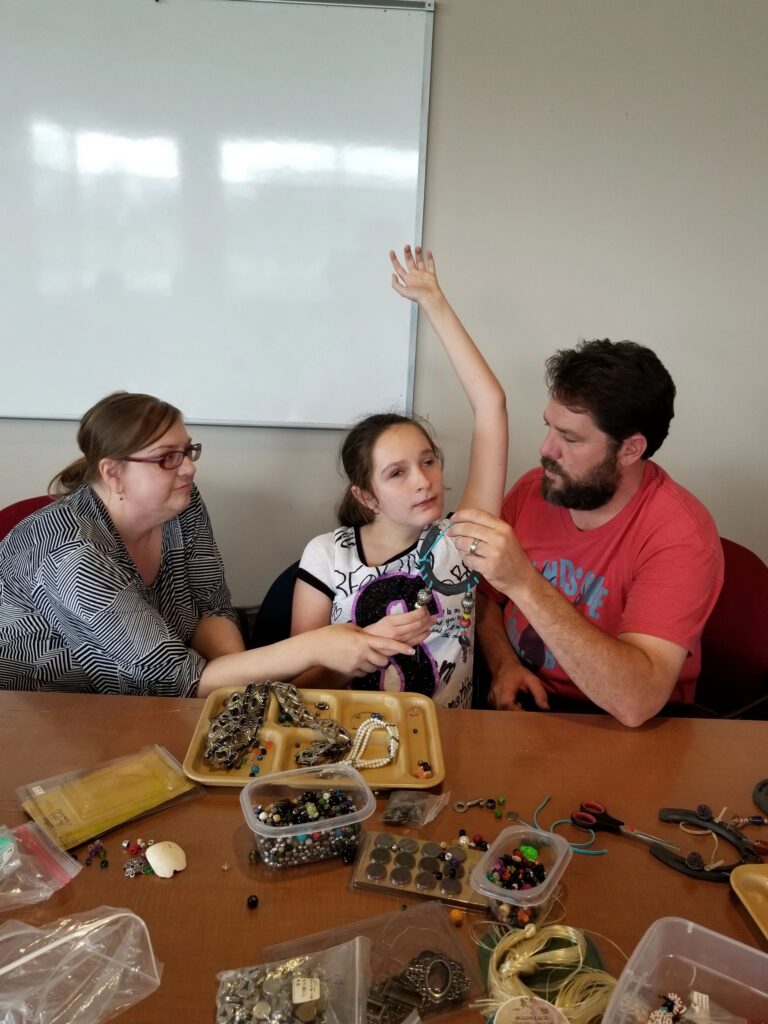 A volunteer helps an artist put on a beaded necklace, trays of beads and charms spread across the table.