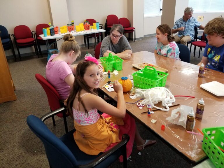 Artists decorate a small pumpkin with paint and glitter glue at a table set with green supply baskets and craft trays.