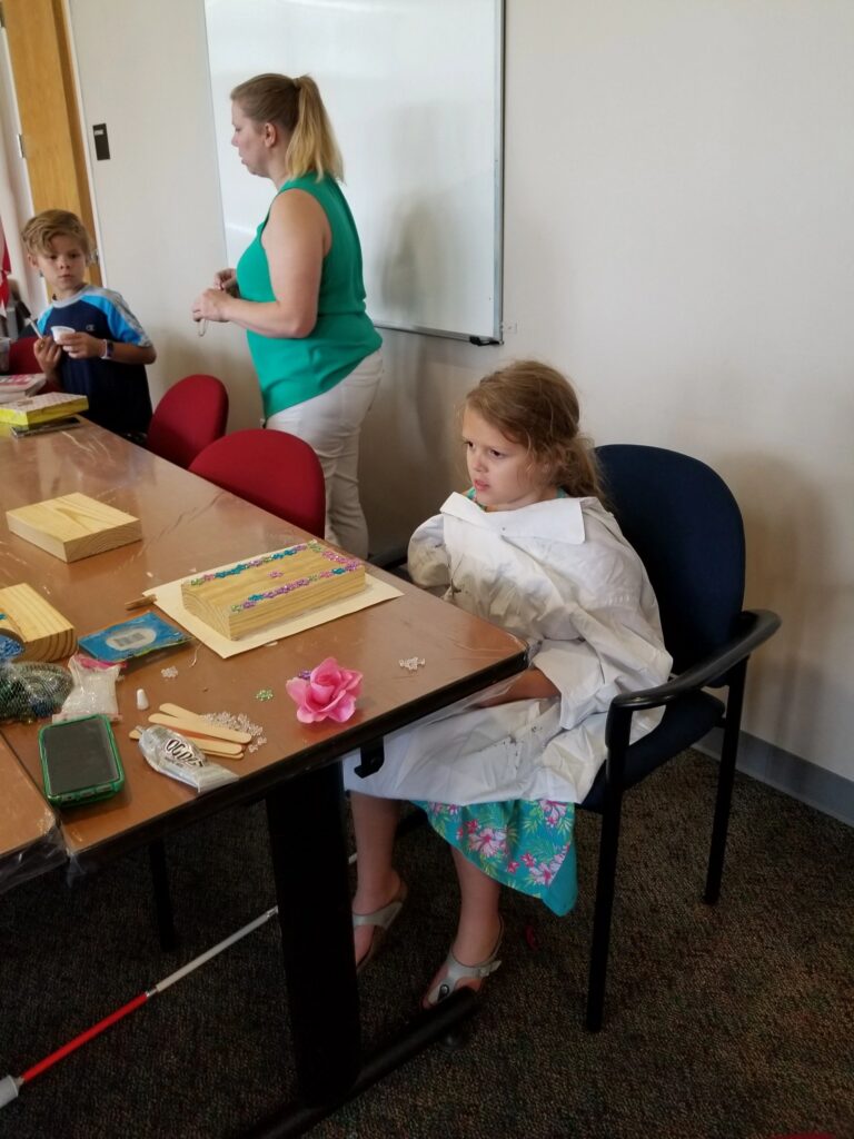 An artist works on a wood block decorated with small flower gems, a pink fabric rose resting nearby on the table.