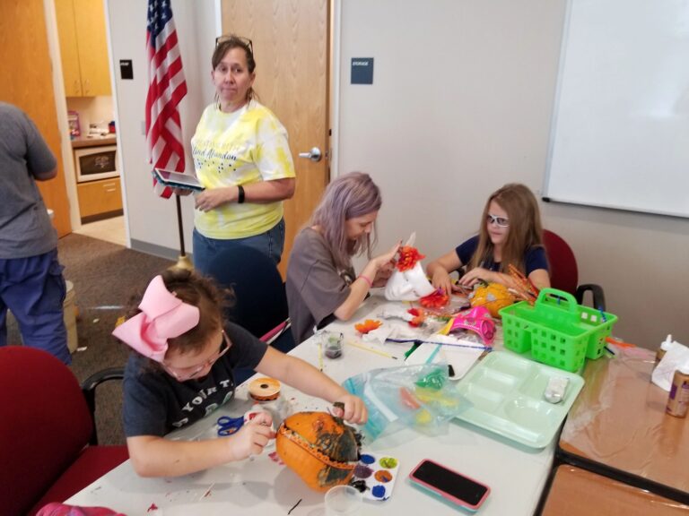 Artists at a table decorate pumpkins with ribbon and ornaments, ranch dressing and craft supplies spread across the surface.