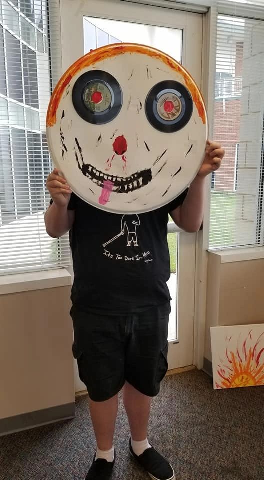An artist holds up a large decorated record painted as a smiling face with two records for eyes, wearing a t-shirt that reads It's Too Dark In Here.