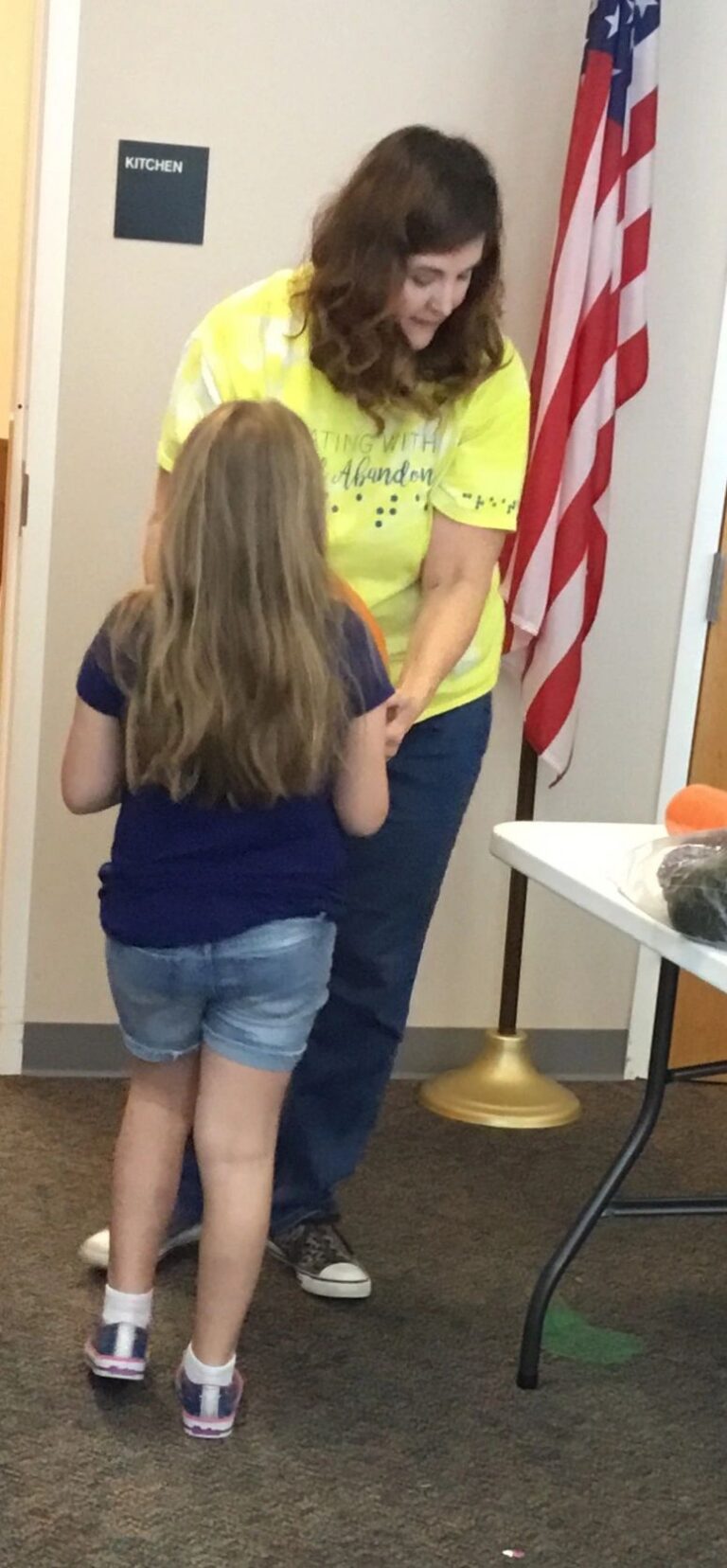 A volunteer walks with an artist near an American flag stand, a small table with craft items behind them.
