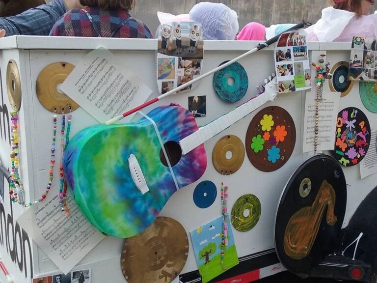 A tie-dye painted acoustic guitar mounted on the float's panel, surrounded by decorated records, a white cane and sheet music.