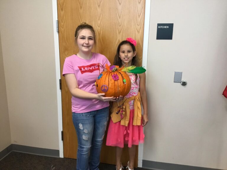 Two artists hold up a pumpkin decorated with a peace sign, painted flowers and feathers around the stem.