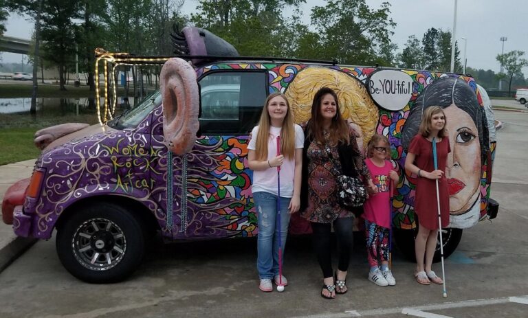 Four people stand in front of the purple Women Rock Art Car, painted with swirling patterns and topped with a large sculpted ear, two holding white canes.