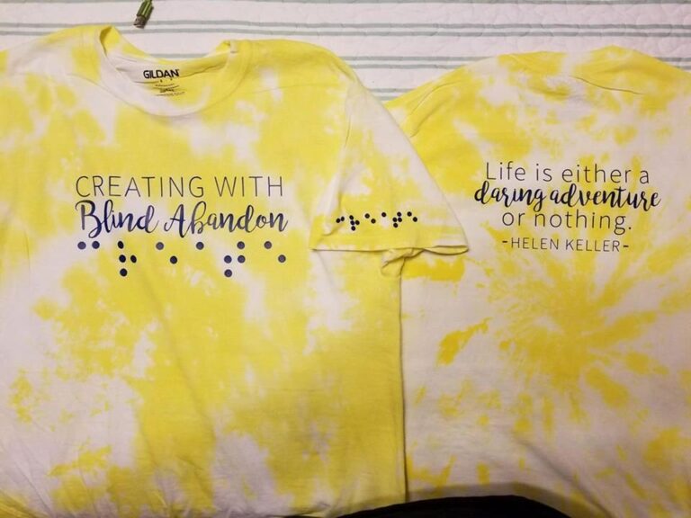 Two folded yellow tie-dye t-shirts, one reading Creating With Blind Abandon with the words spelled out in raised braille dots on the sleeve, the other printed with a Helen Keller quote.