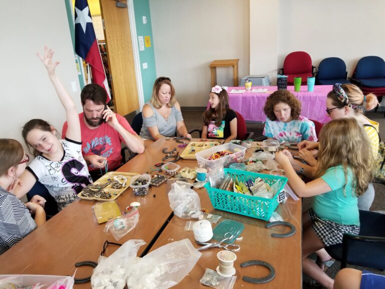 A large group of artists and volunteers sit around a table decorating horseshoes with beads, trays of small charms spread across the surface.