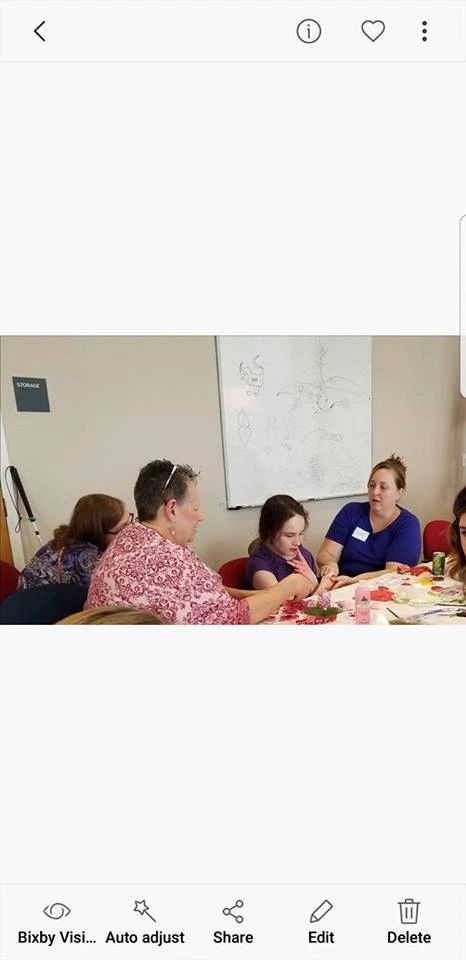 A photo shown on a phone screen of artists and a volunteer seated at a table, working on a craft project in front of a whiteboard sketched with a dragon.