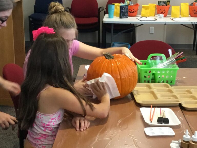 Two artists wipe down a pumpkin with a cloth at a table set with paint trays and a green supply basket.