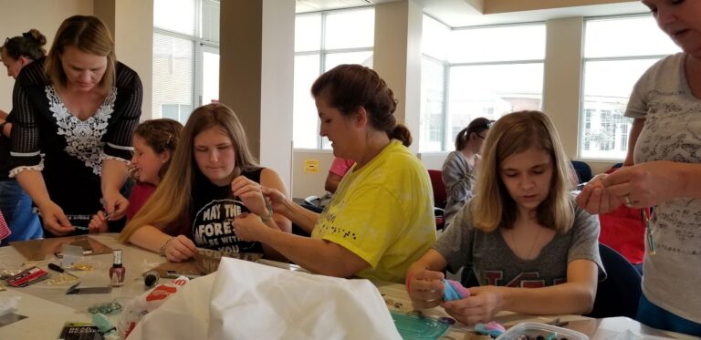 Several artists and a volunteer bead together at a table, small trays of beads and nail polish scattered across the surface.