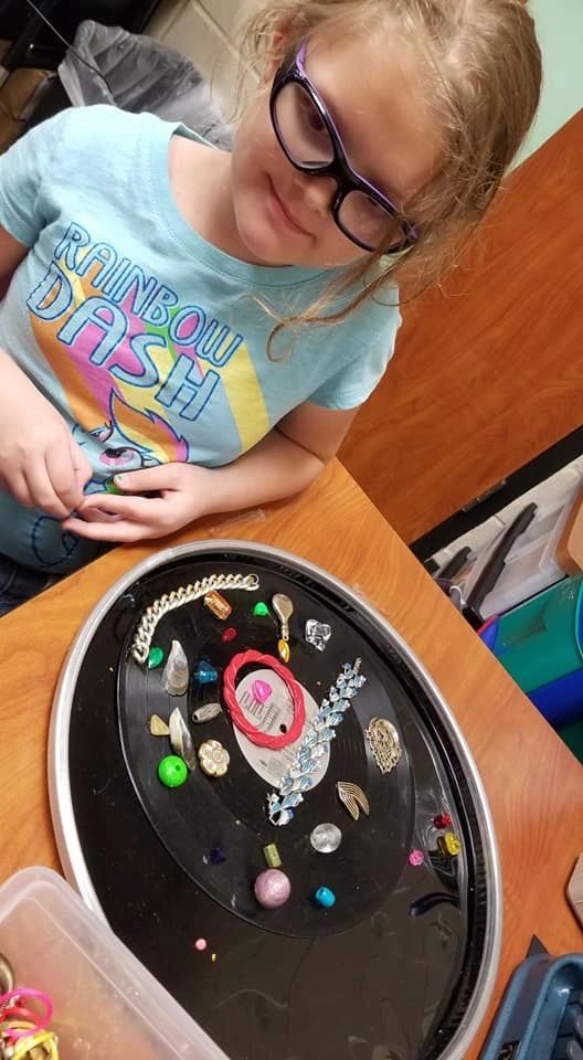 An artist arranges small charms and beads on a decorated record, a beaded bracelet and pendant visible among the pieces.