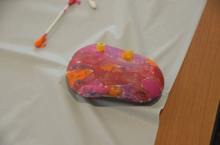 A stone painted pink and orange with two small yellow beads pressed into the surface, resting on a table beside a cotton swab.