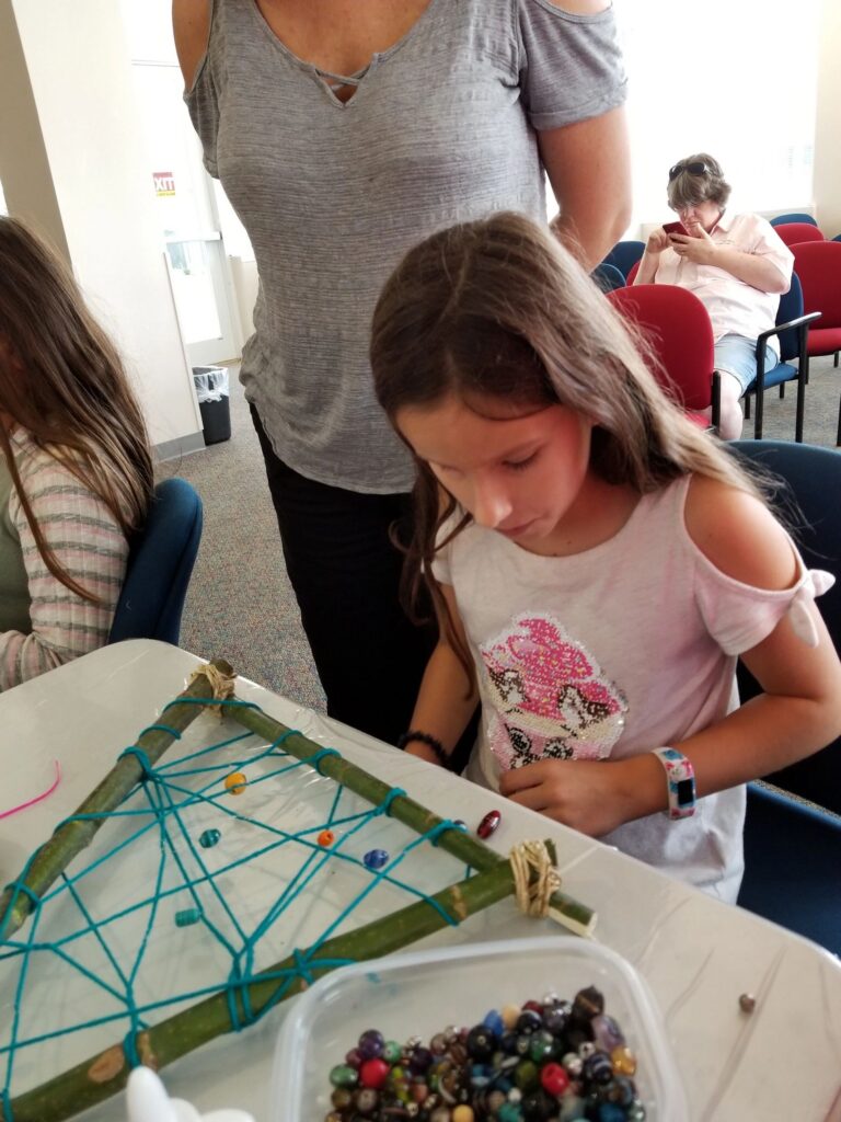 A volunteer stands beside an artist weaving beads onto a stick-frame dream catcher, a bin of beads on the table.