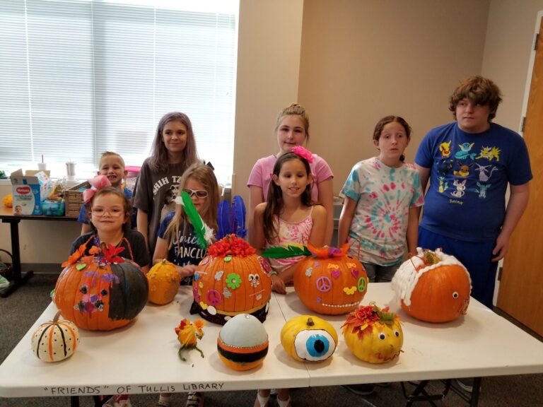 Eight artists pose together behind a table lined with decorated pumpkins in a range of designs, from googly-eyed monsters to peace-sign florals.