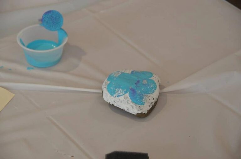 A stone painted purple with pink, red and blue paint dabbed over the surface, resting on a table splattered with purple paint.
