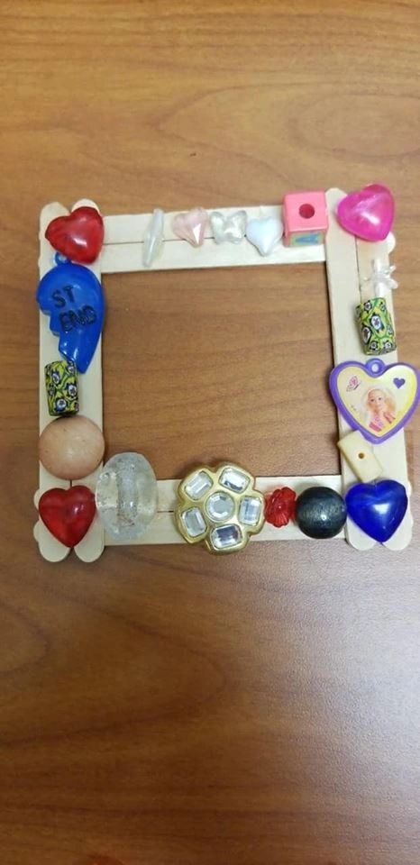A small picture frame made from craft sticks, decorated with heart charms, a framed photo, glass gems and a beaded flower cluster.