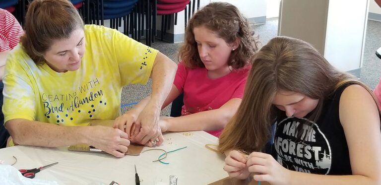 A volunteer helps an artist bead on a clipboard while another artist works nearby, cord and small tools spread on the table.