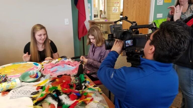 Artists sit at a table covered in feathers and craft supplies while a videographer films them working.