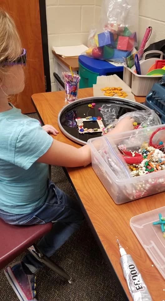 An artist works on a small picture frame with beads and charms, a bin of mixed beads and jewellery nearby on the table.