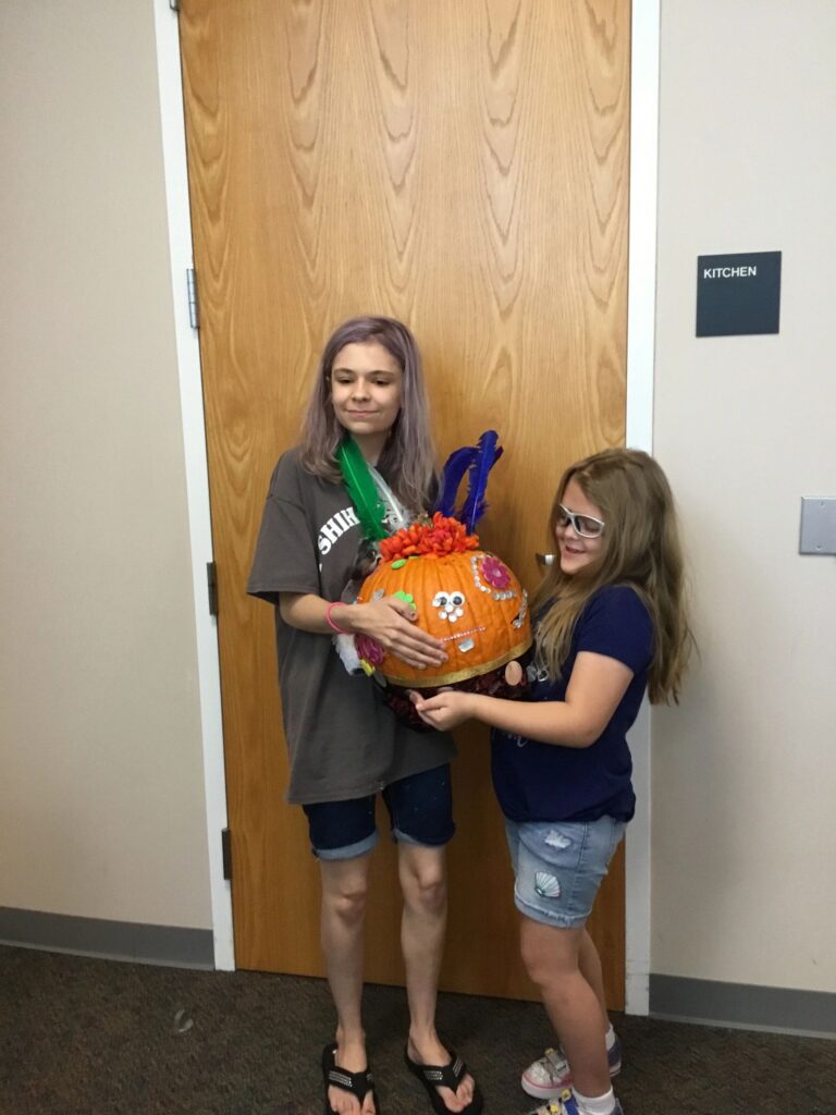 Two artists hold up a pumpkin covered in painted flowers and a face made from googly eyes, topped with green and blue feathers.