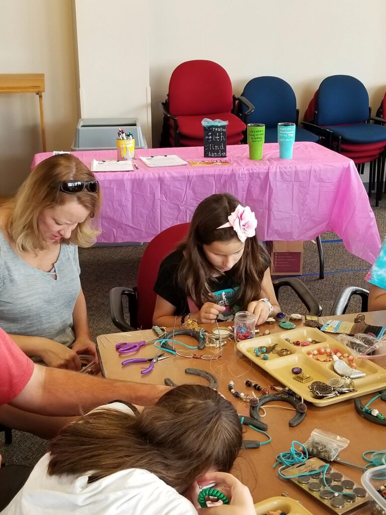 A volunteer helps two artists decorate horseshoes with beads at a table set with trays of charms and small trinkets.