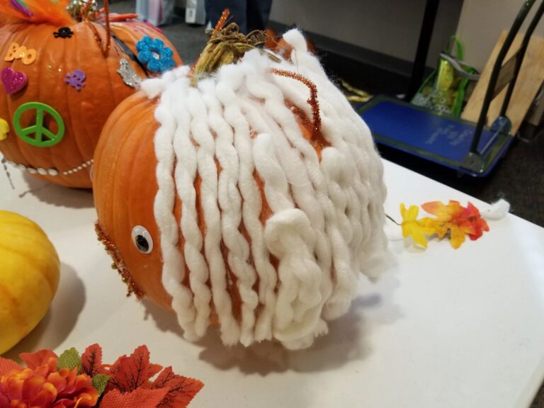 A pumpkin wrapped in white yarn with a single googly eye and rust-coloured pipe cleaner accents, sitting beside other decorated pumpkins.