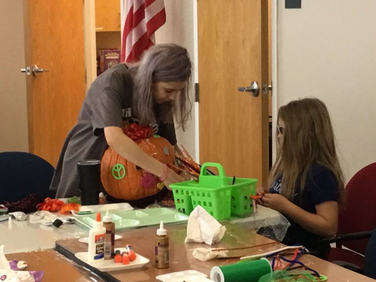 An artist lifts a decorated pumpkin with orange flowers and a peace sign onto a table, a green basket nearby.