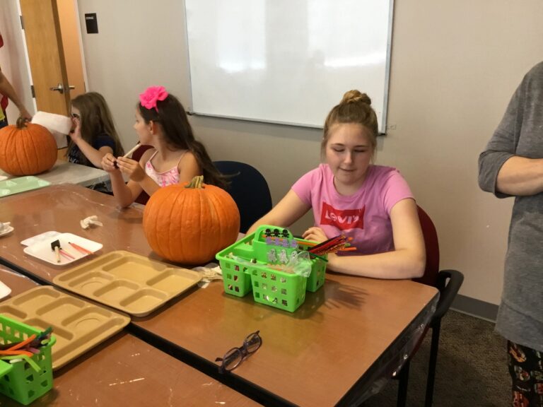 An artist sits at a table with a carved-face pencil cup and a whole pumpkin, colourful pencils nearby.