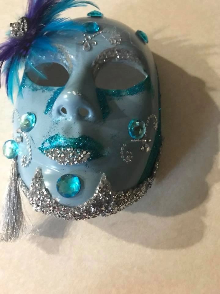 A decorated masquerade mask in pale blue, glitter and rhinestones, with teal glitter accents around the eyes and lips and a tassel hanging from one side.