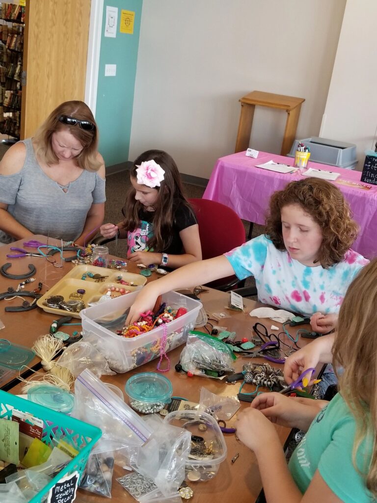 Two artists and a volunteer sort through bins of beads and charms at a table set with horseshoes and small trays.