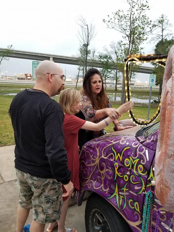 Three people gather around the front of the Women Rock Art Car, one reaching up to touch its painted hood lettering, with floodwater and an overpass in the background.