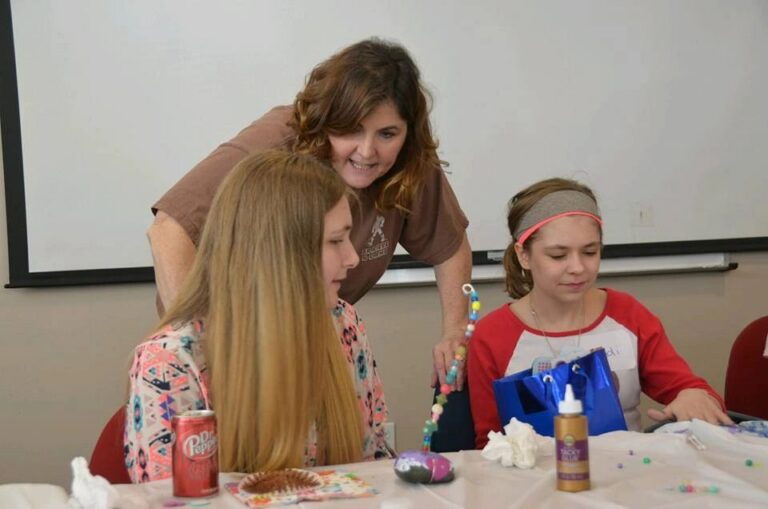 A volunteer helps two artists with a beaded craft at a table, one artist working on a beaded strand.