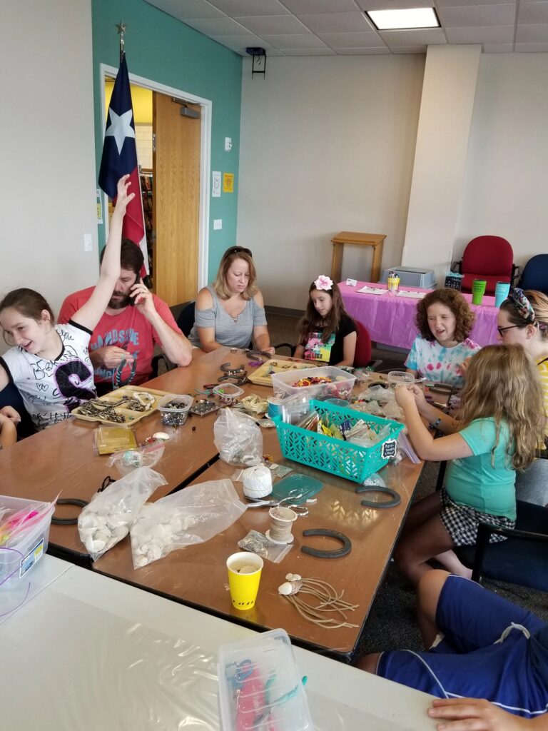 A large group of artists and volunteers sit around a table decorating horseshoes with beads and charms, a Texas flag in the corner.