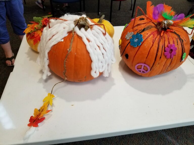 Two decorated pumpkins on a table: one wrapped in white yarn with twine trailing down, the other splattered with black paint and topped with colourful feathers.