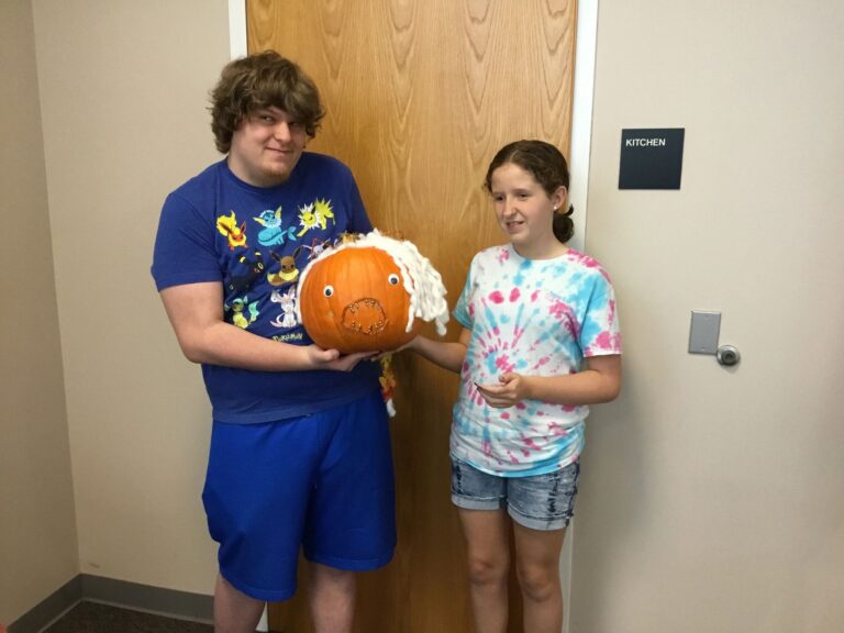 Two artists hold up a pumpkin wrapped in white yarn with googly eyes and gold-studded accents.