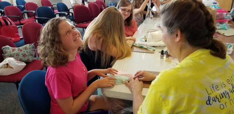 A volunteer helps an artist with a beaded bracelet at a table set with fabric pouches and craft supplies, a Helen Keller quote visible on another artist's shirt nearby.