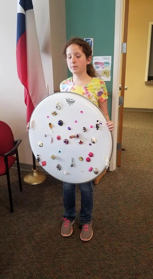 An artist holds up a large drumhead dotted with dozens of small pins, charms and gems arranged across its surface.