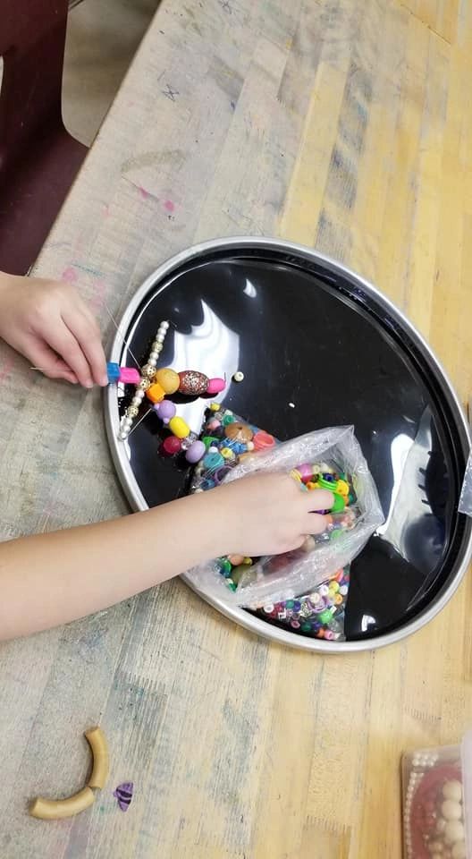 A hand threads beads onto a wire figure, with a bag of loose beads nearby on a wooden table.