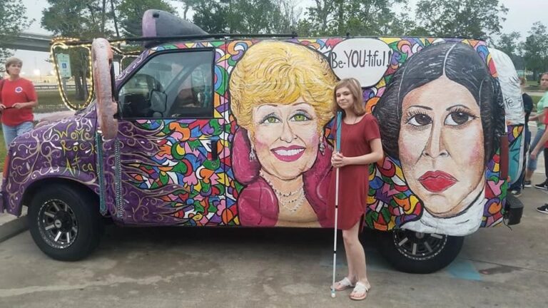 Two people stand before the Women Rock Art Car, painted with portraits of two women and a Be-YOU-tiful speech bubble, one holding a white cane.