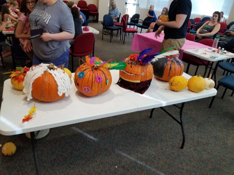 Two artists hold up a pumpkin decorated with painted flowers, feathers around the stem, and a face made from googly eyes and glitter pipe cleaners.