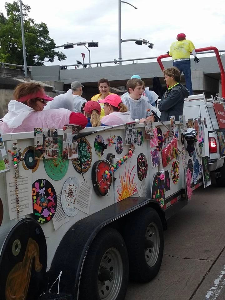 Riders on the float pass beneath an overpass, several wearing rain ponchos, with decorated records and painted panels on display beside them.