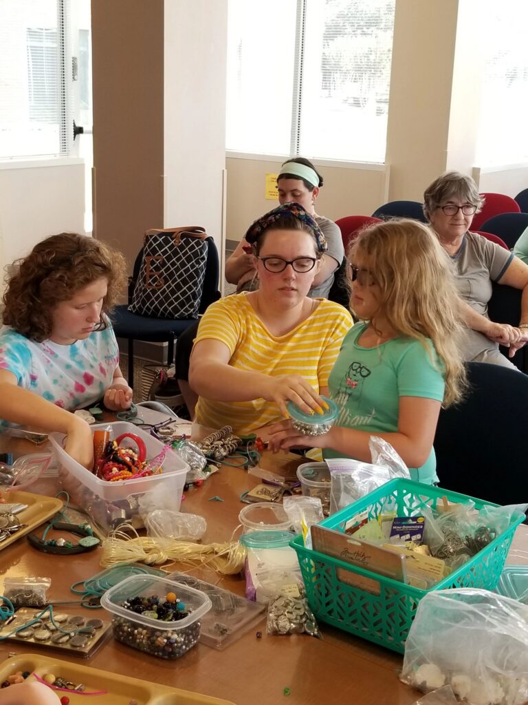 A volunteer helps two artists decorate horseshoes at a table covered in bead containers, raffia and small trinkets.