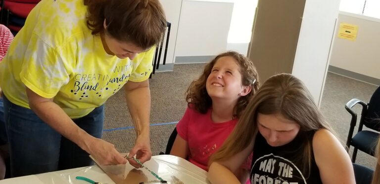 A volunteer helps an artist bead at the table while another artist smiles beside her.