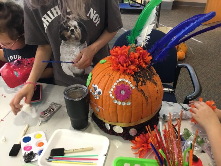 A pumpkin decorated with a pink flower gem, googly eyes and rhinestone trim, topped with orange mums and green, white and blue feathers.